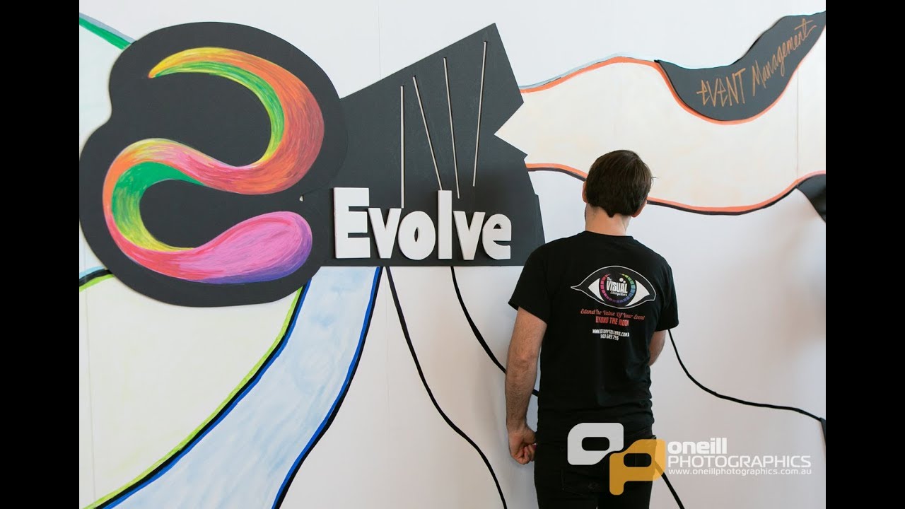 Person standing in front of colorful mural with "Evolve" text, showcasing visual storytelling at the Evolve Conference by The Visual Storytellers Group.