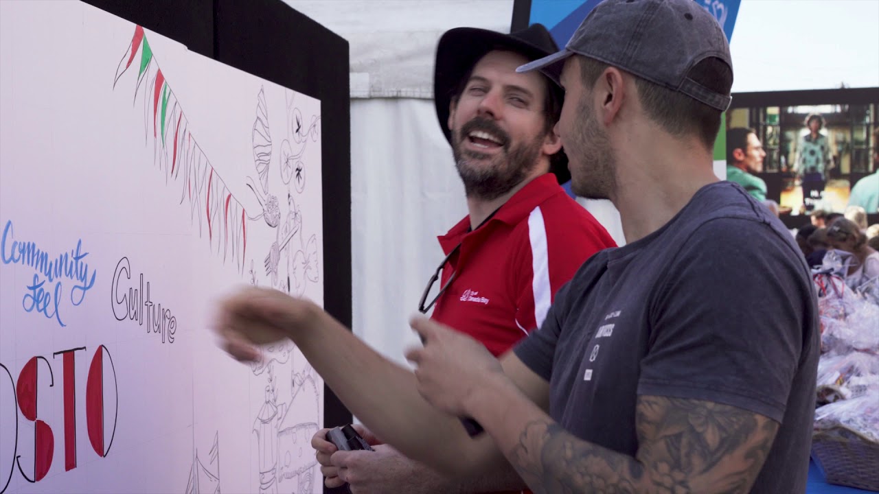 Two men engaging in conversation while creating a live interactive mural at a festival, featuring the words "Community," "Feel," and "Culture" on the mural, emphasizing visual storytelling and audience engagement.