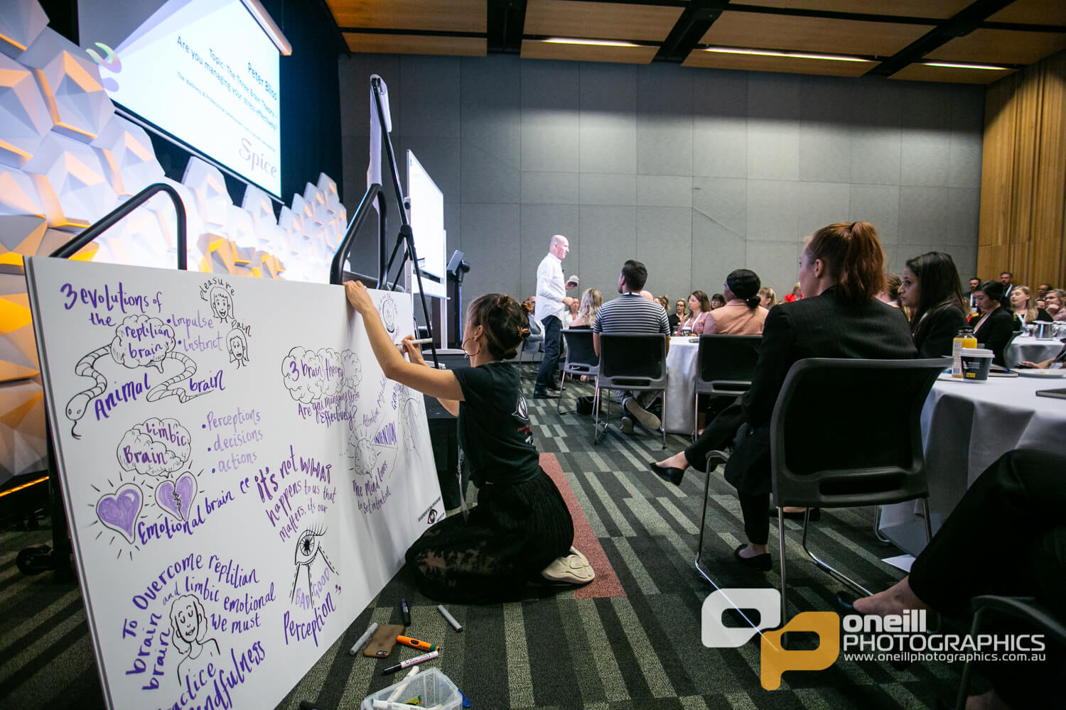 Participant illustrating key concepts on a whiteboard during a workshop on visual storytelling and communication, with an audience engaged in the background.