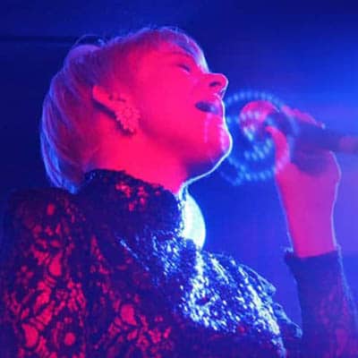 Emma Deans, Head Communications Trainer, performing passionately on stage with a microphone, illuminated by vibrant blue and purple lights.