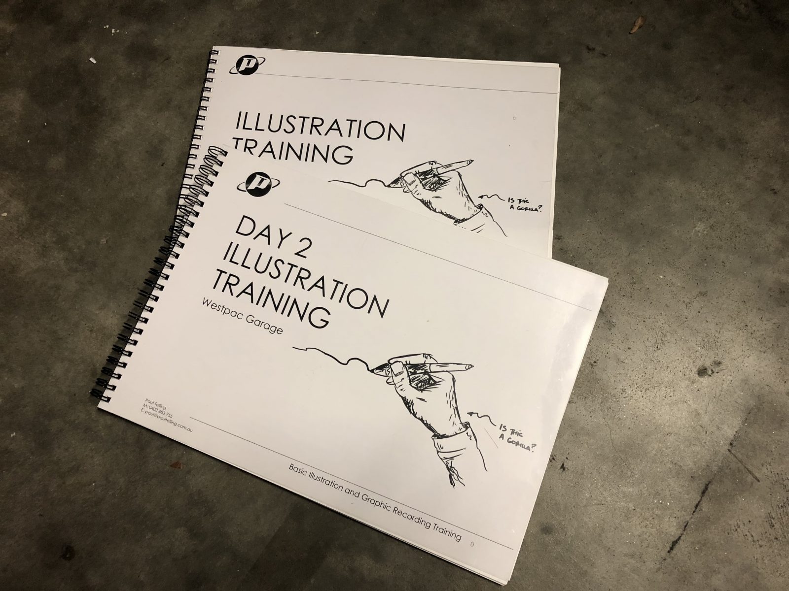 Illustration training manuals stacked on a concrete surface, featuring a hand holding a pen with a sketch and text asking, "Is this a gorilla?"