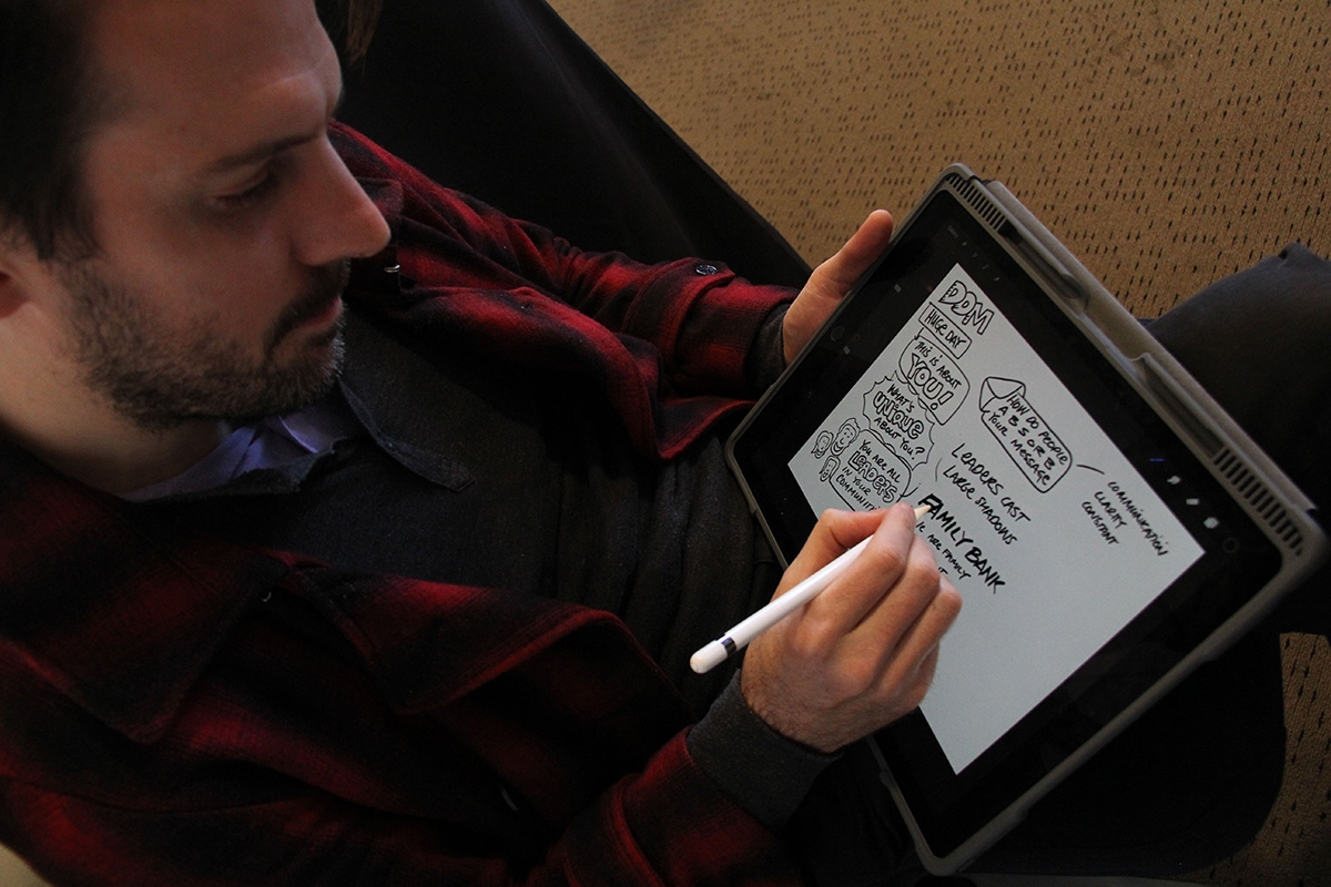 A man in a red and black plaid jacket drawing on a tablet with a stylus, creating a sketched diagram with text and illustrations